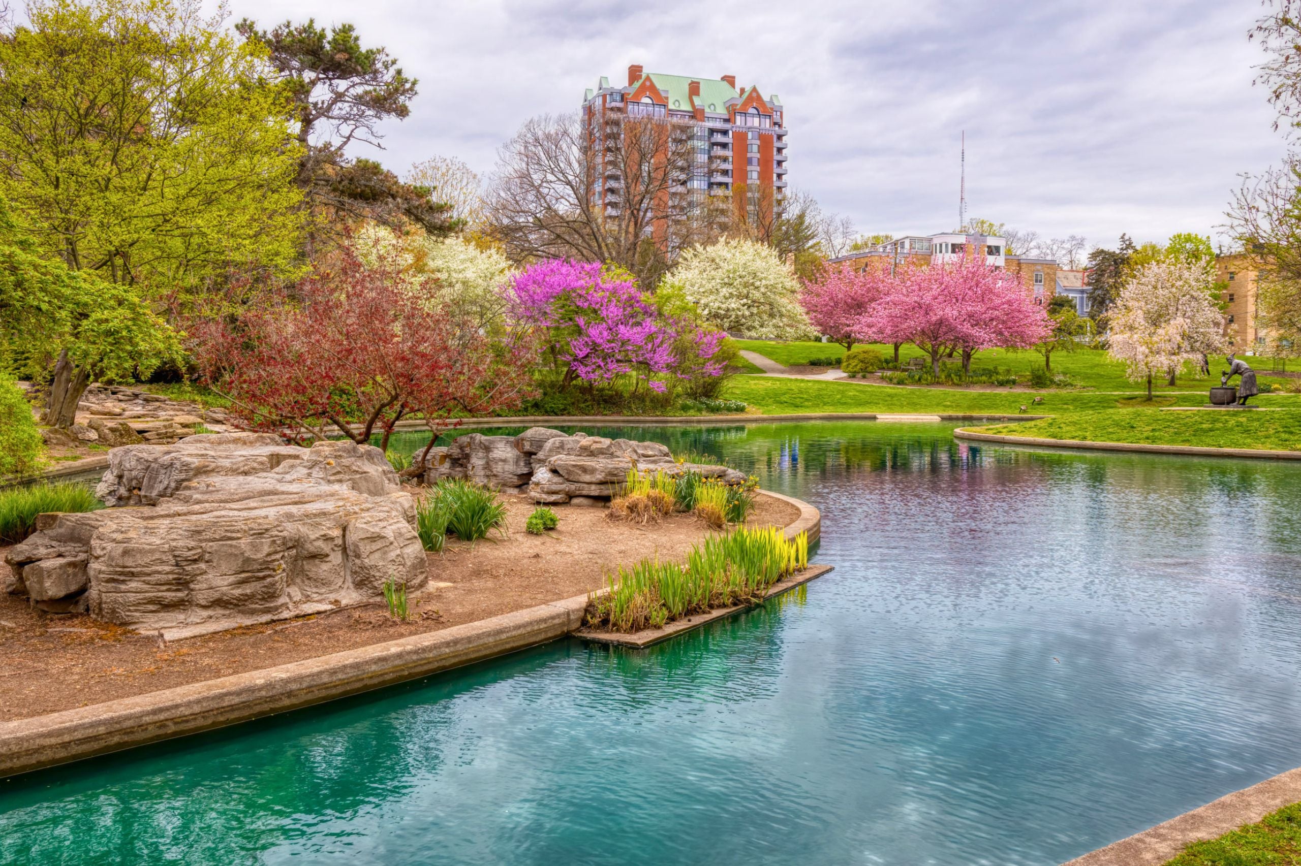 Eden Park in spring with a curved turquoise pond, large rocks, and blooming pink, white, and purple trees under a cloudy sky. A tall residential building rises in the background beyond the green lawns and walking paths.
