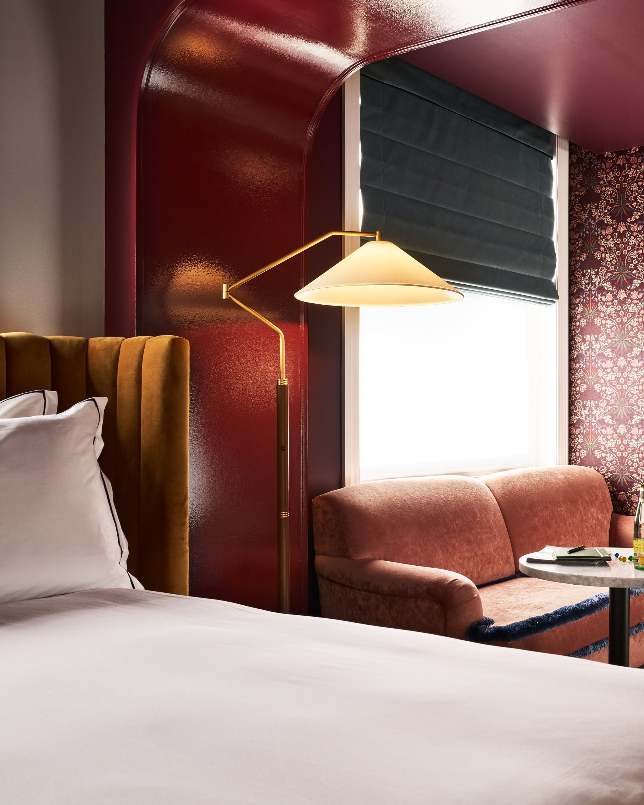 A brass floor lamp with a conical shade illuminates a richly decorated bedroom. A mustard bed and pink velvet couch sit beside deep red walls with patterned wallpaper.