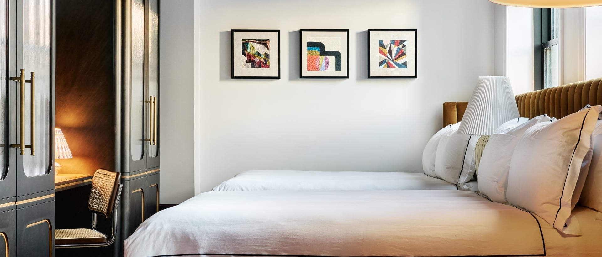 A large bed with white bedding and a brown headboard fills a modern bedroom. A gold chandelier hangs from the dark purple ceiling, flanked by dark cabinets and abstract art.