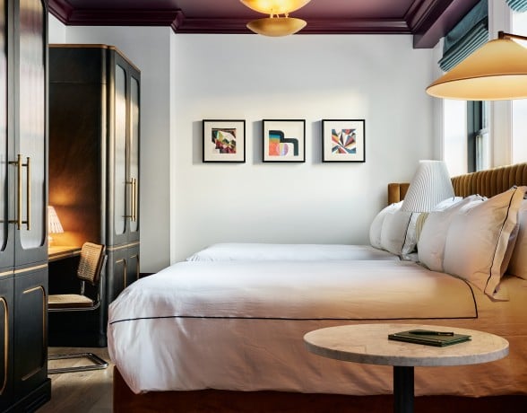 A large bed with white bedding and a brown headboard fills a modern bedroom. A gold chandelier hangs from the dark purple ceiling, flanked by dark cabinets and abstract art.
