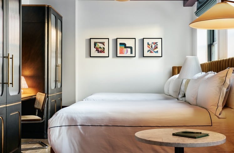 A large bed with white bedding and a brown headboard fills a modern bedroom. A gold chandelier hangs from the dark purple ceiling, flanked by dark cabinets and abstract art.
