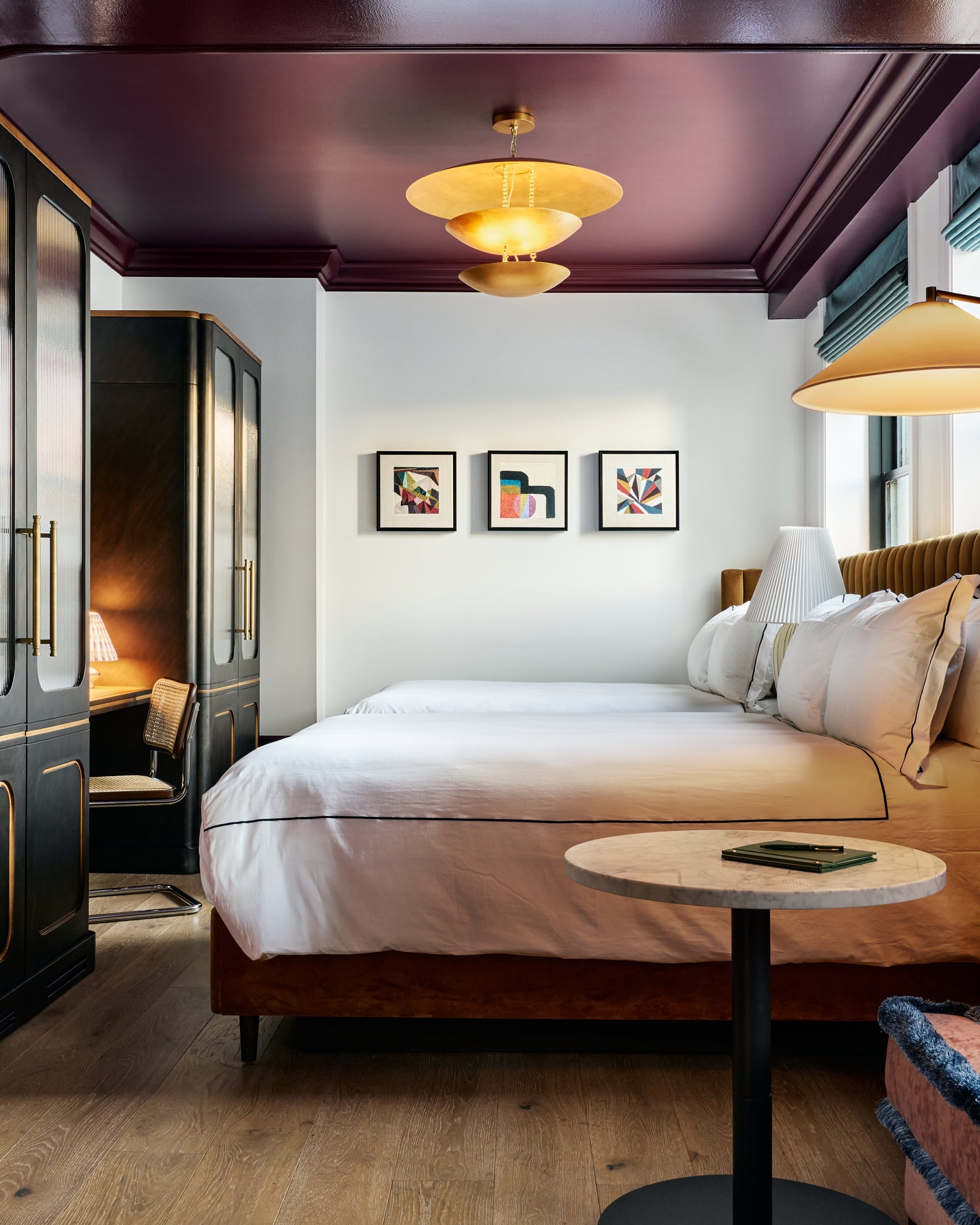 A large bed with white bedding and a brown headboard fills a modern bedroom. A gold chandelier hangs from the dark purple ceiling, flanked by dark cabinets and abstract art.
