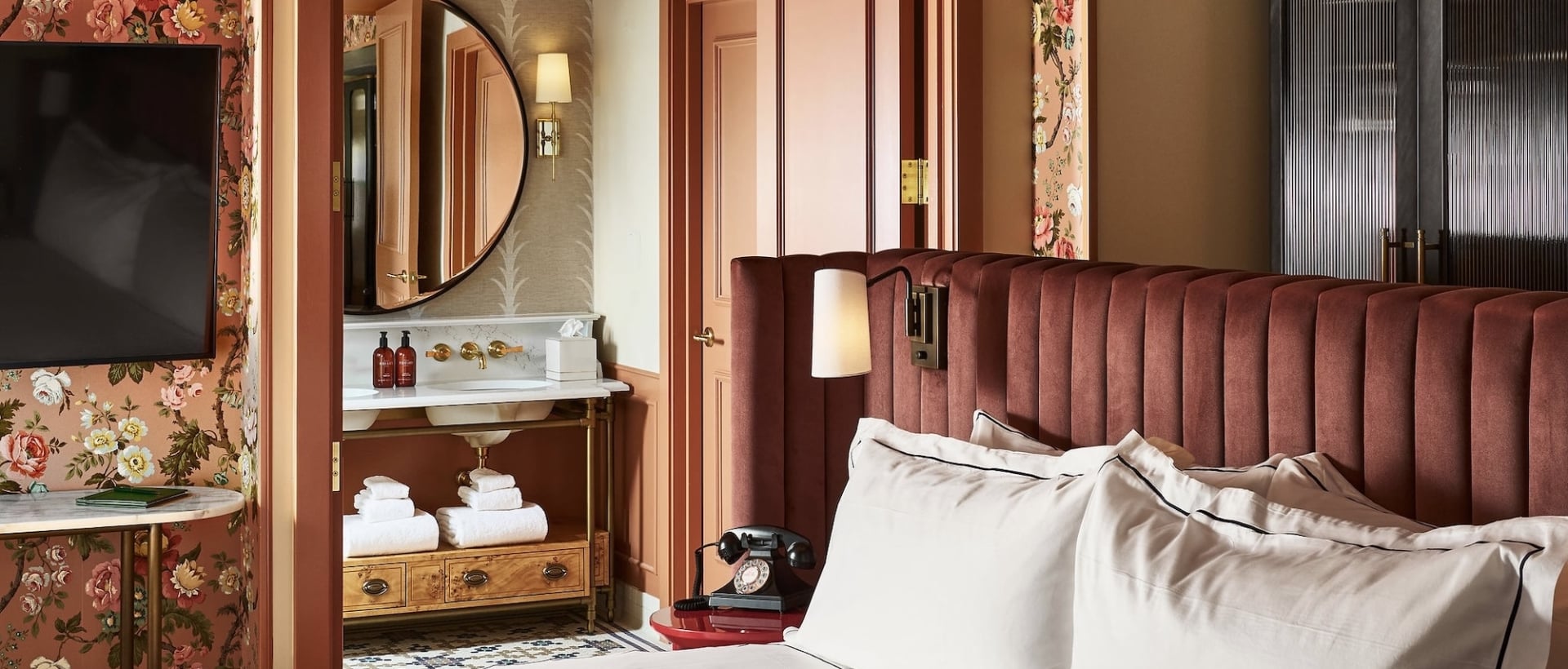 White-linened bed with dark red velvet headboard. Lavish room features floral wallpaper, vanity with mirror and two bottles labeled MALIN+GOETZ, plus a rotary phone on a red side table.