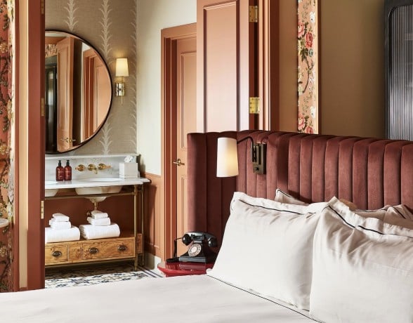 White-linened bed with dark red velvet headboard. Lavish room features floral wallpaper, vanity with mirror and two bottles labeled MALIN+GOETZ, plus a rotary phone on a red side table.