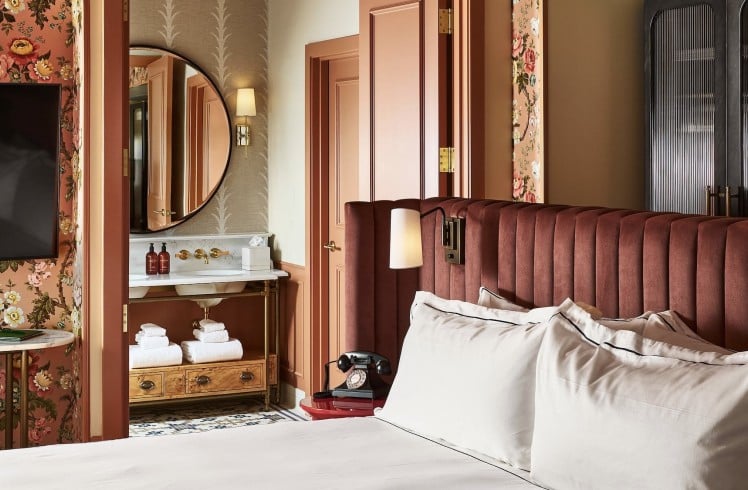 White-linened bed with dark red velvet headboard. Lavish room features floral wallpaper, vanity with mirror and two bottles labeled MALIN+GOETZ, plus a rotary phone on a red side table.