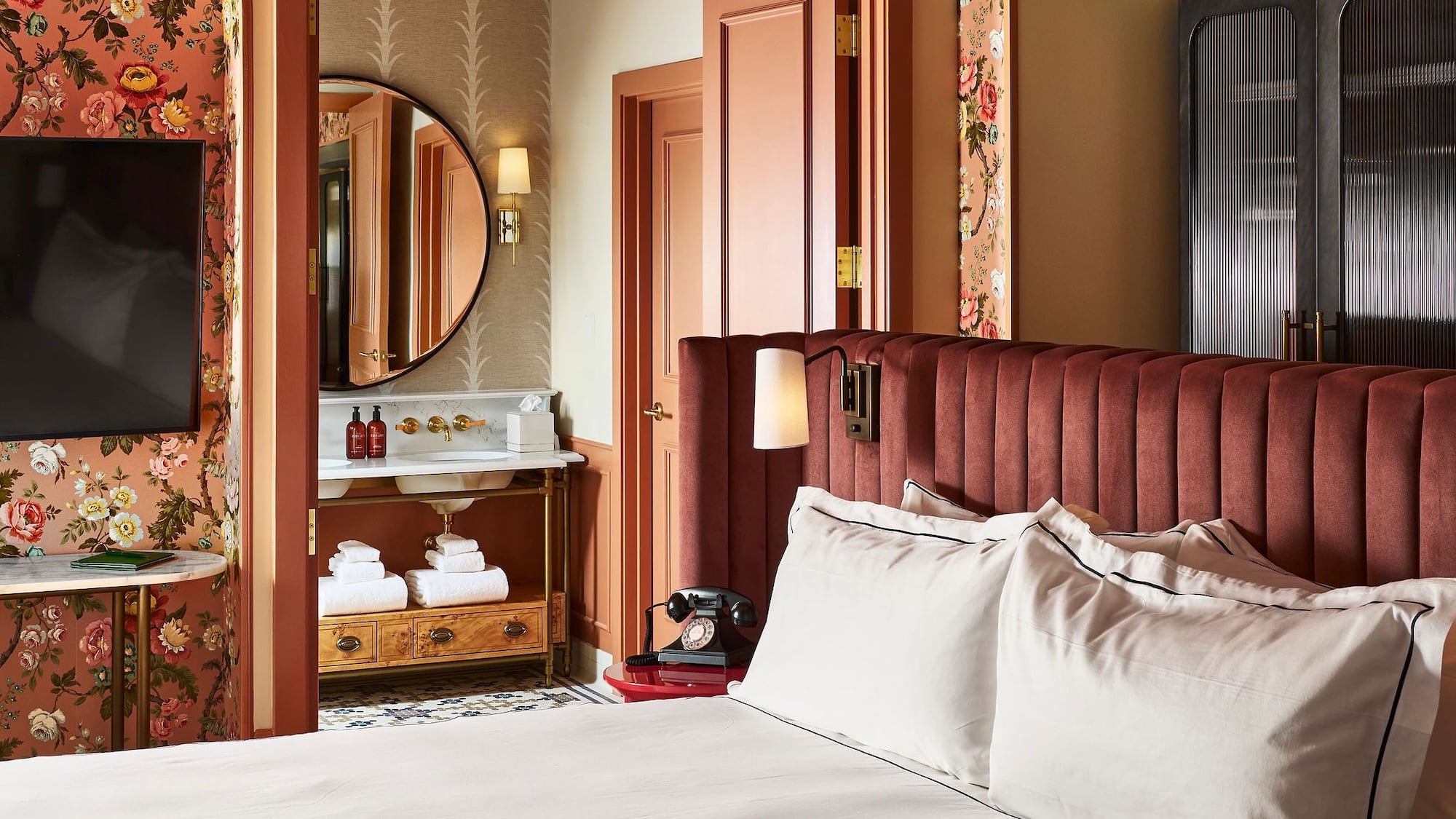 White-linened bed with dark red velvet headboard. Lavish room features floral wallpaper, vanity with mirror and two bottles labeled MALIN+GOETZ, plus a rotary phone on a red side table.