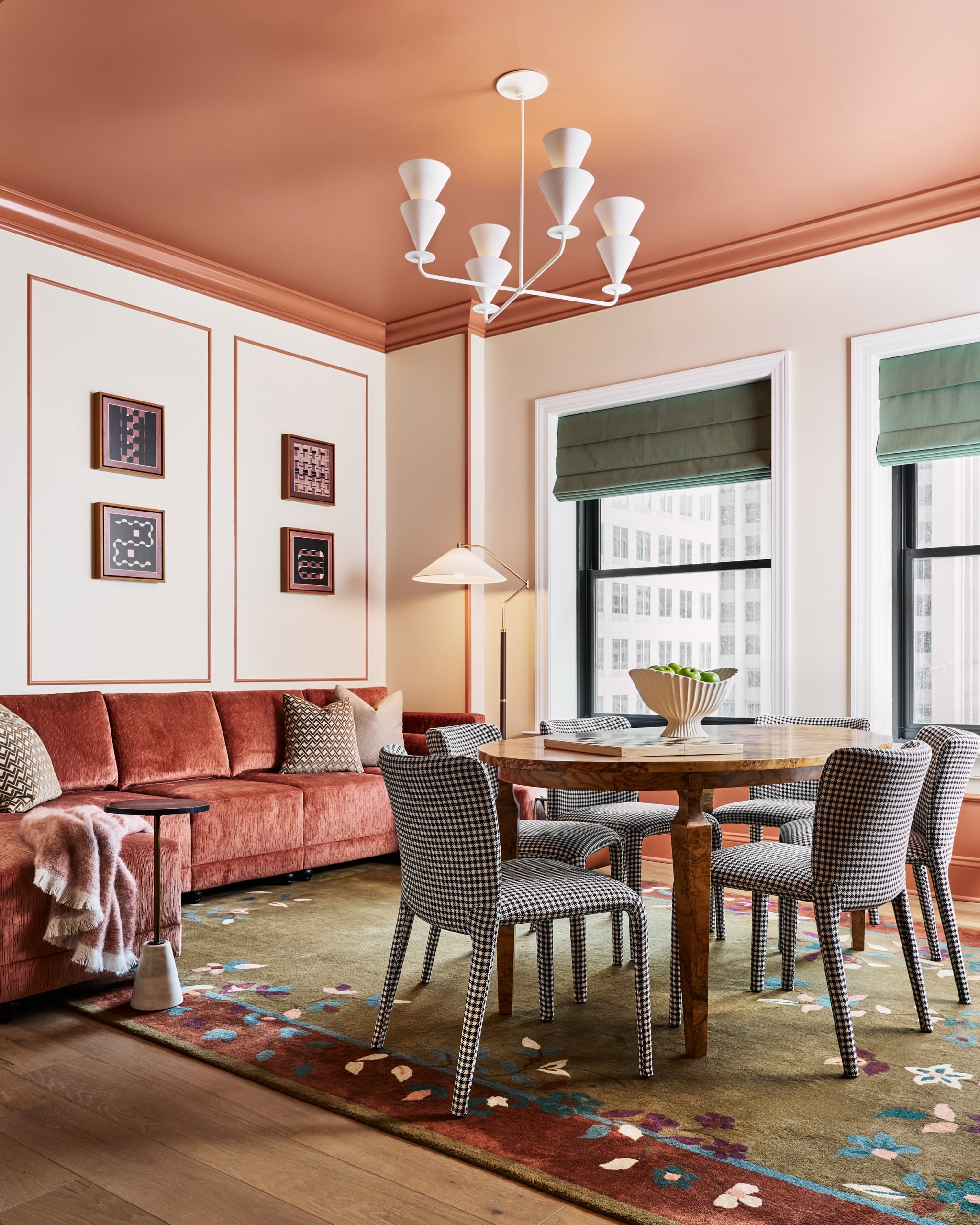 A contemporary room features a rust sectional sofa, a round wooden dining table with houndstooth chairs, and a floral rug. Salmon-pink ceiling and city view windows frame the space.