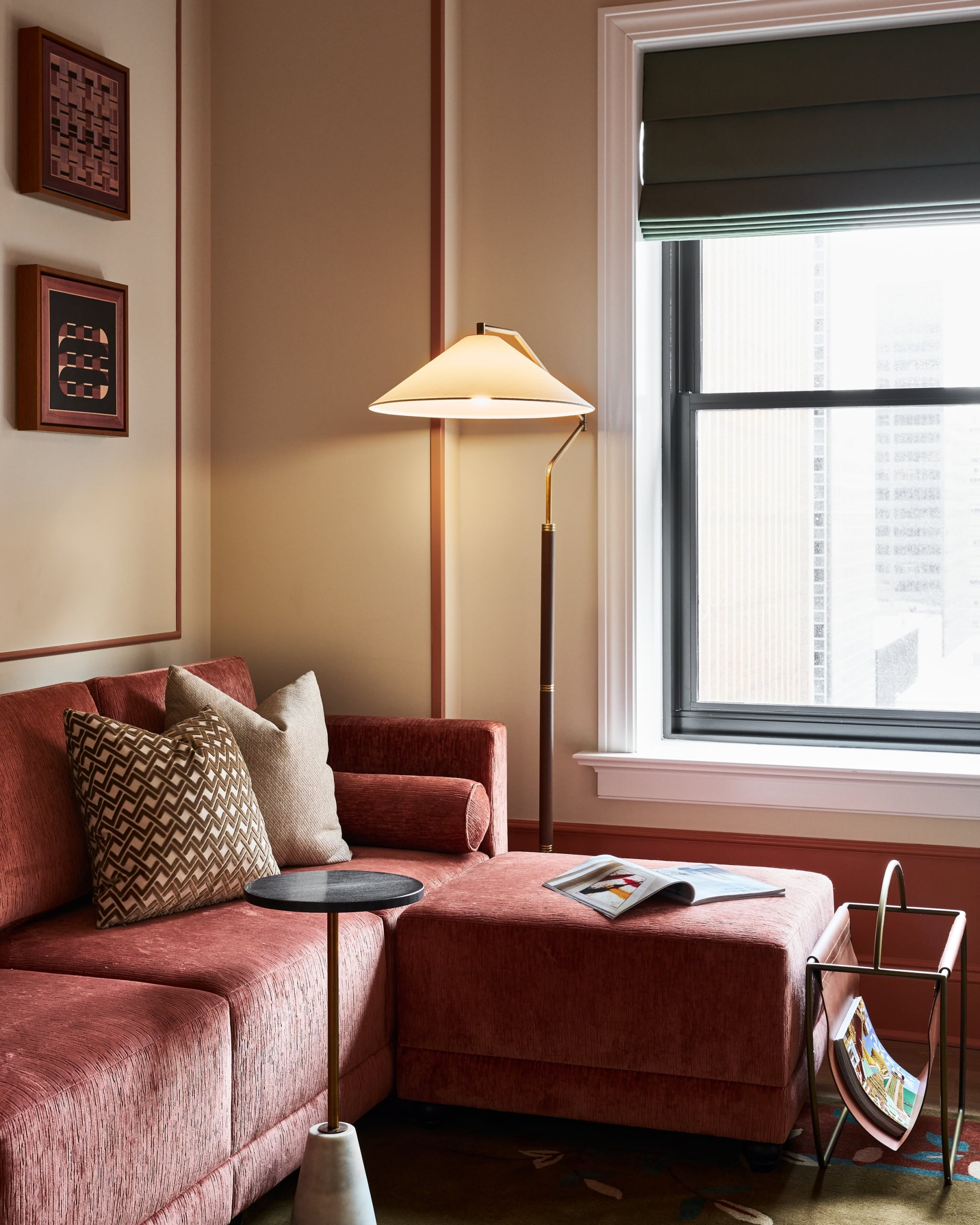 A burnt-orange L-shaped sofa with pillows dominates a cozy room. A lit floor lamp and side table sit by a large window overlooking city buildings. Wall art decorates beige walls.