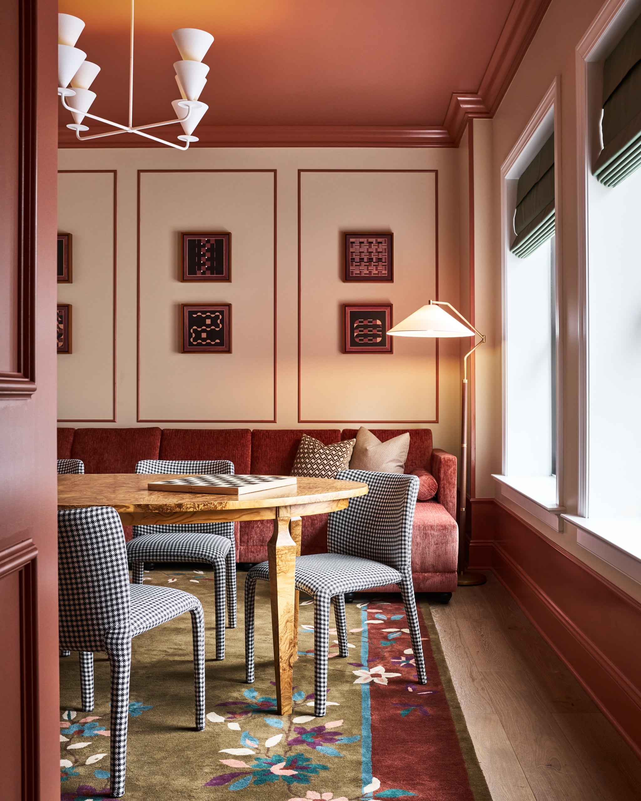 A game table holds a chessboard, surrounded by houndstooth chairs. The room features a red sofa, abstract art, a floor lamp, a floral rug, and rust-red and beige walls.