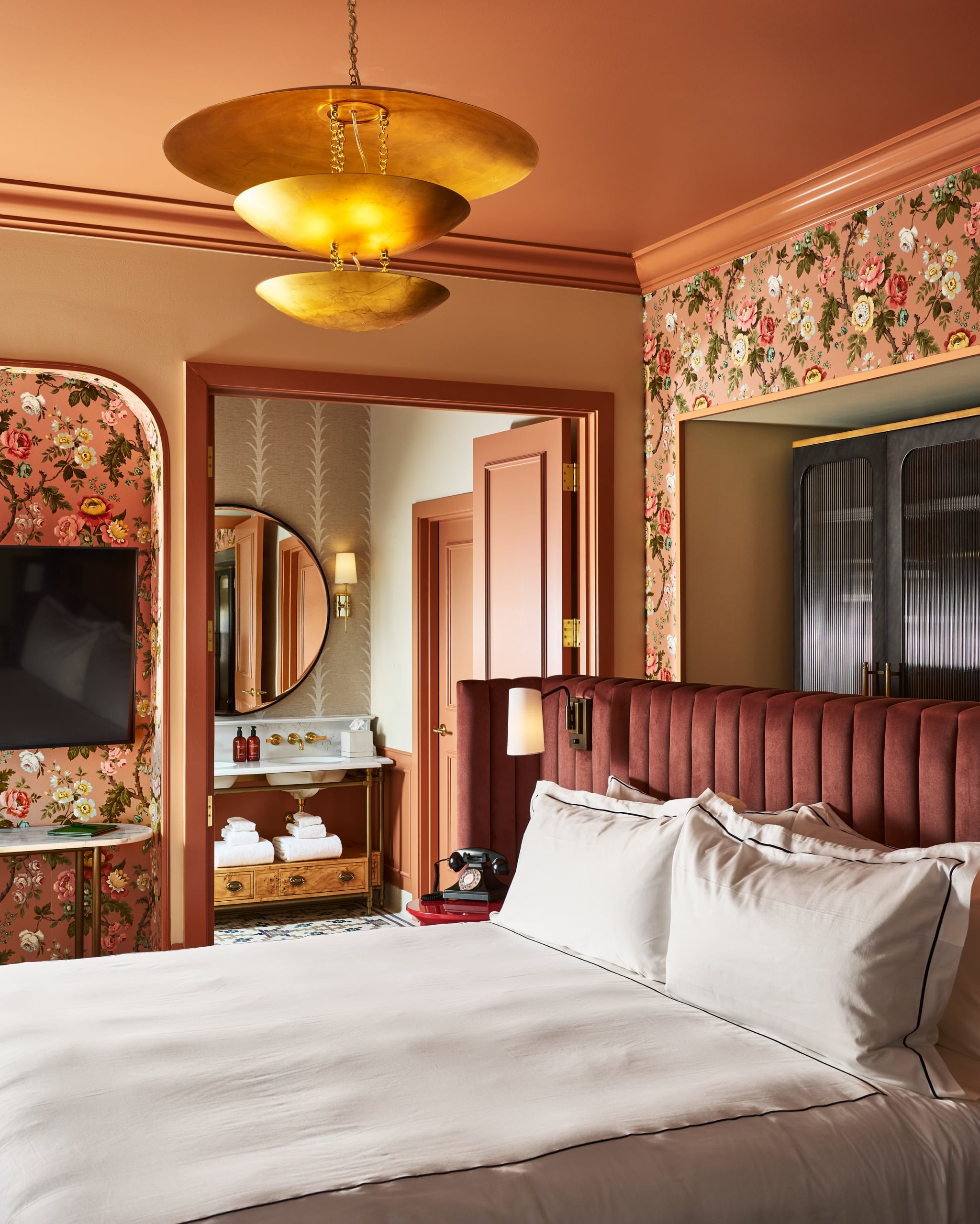 A plush bed with a maroon headboard fills the foreground of a hotel room. Floral walls, a gold chandelier, and a bathroom with a sink and mirror are visible.