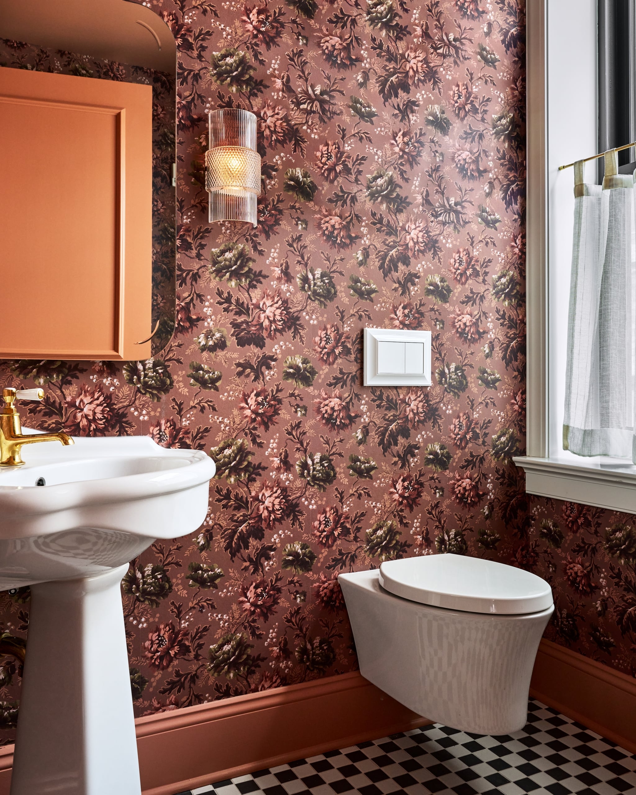 Pedestal sink and wall-mounted toilet in a bathroom with dark floral wallpaper. A peach cabinet, ribbed sconce, and checkered floor tiles complete the vivid interior.