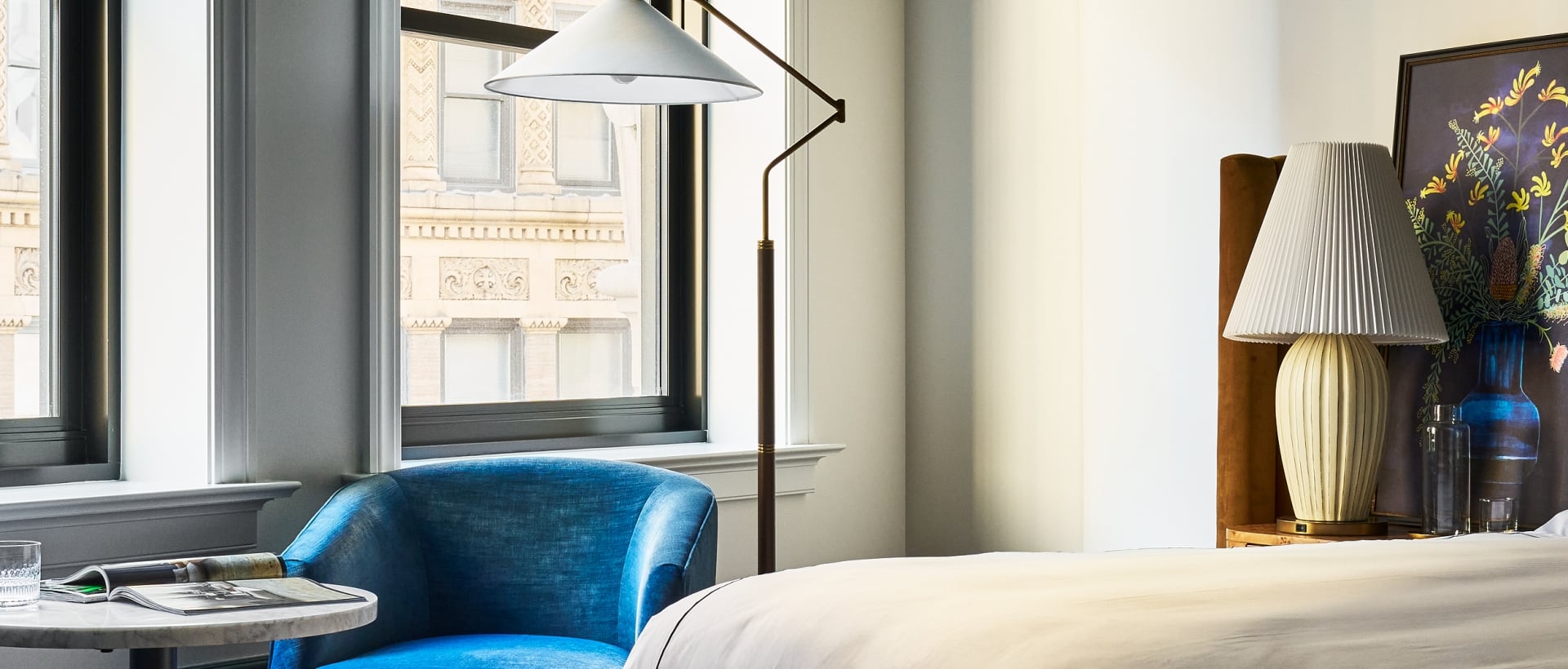 A plush blue armchair sits by tall windows overlooking ornate city buildings. A bed with white sheets and a patterned rug furnish the elegant room.