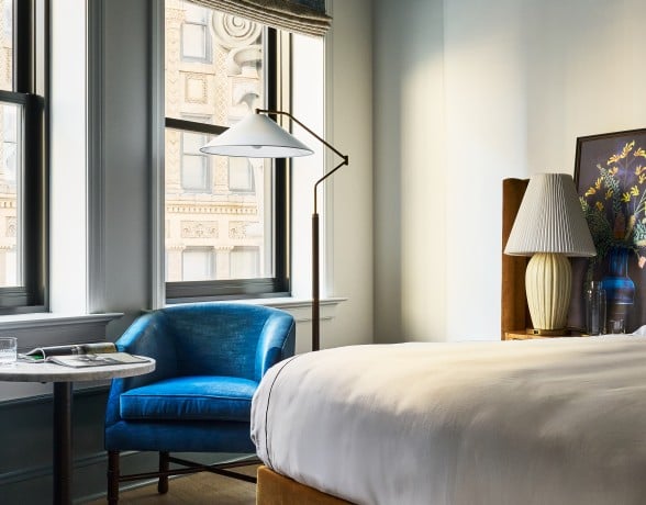 A plush blue armchair sits by tall windows overlooking ornate city buildings. A bed with white sheets and a patterned rug furnish the elegant room.