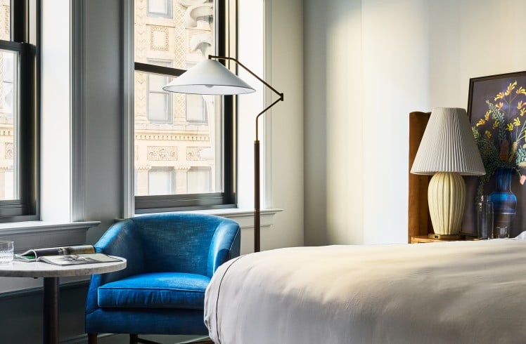 A plush blue armchair sits by tall windows overlooking ornate city buildings. A bed with white sheets and a patterned rug furnish the elegant room.