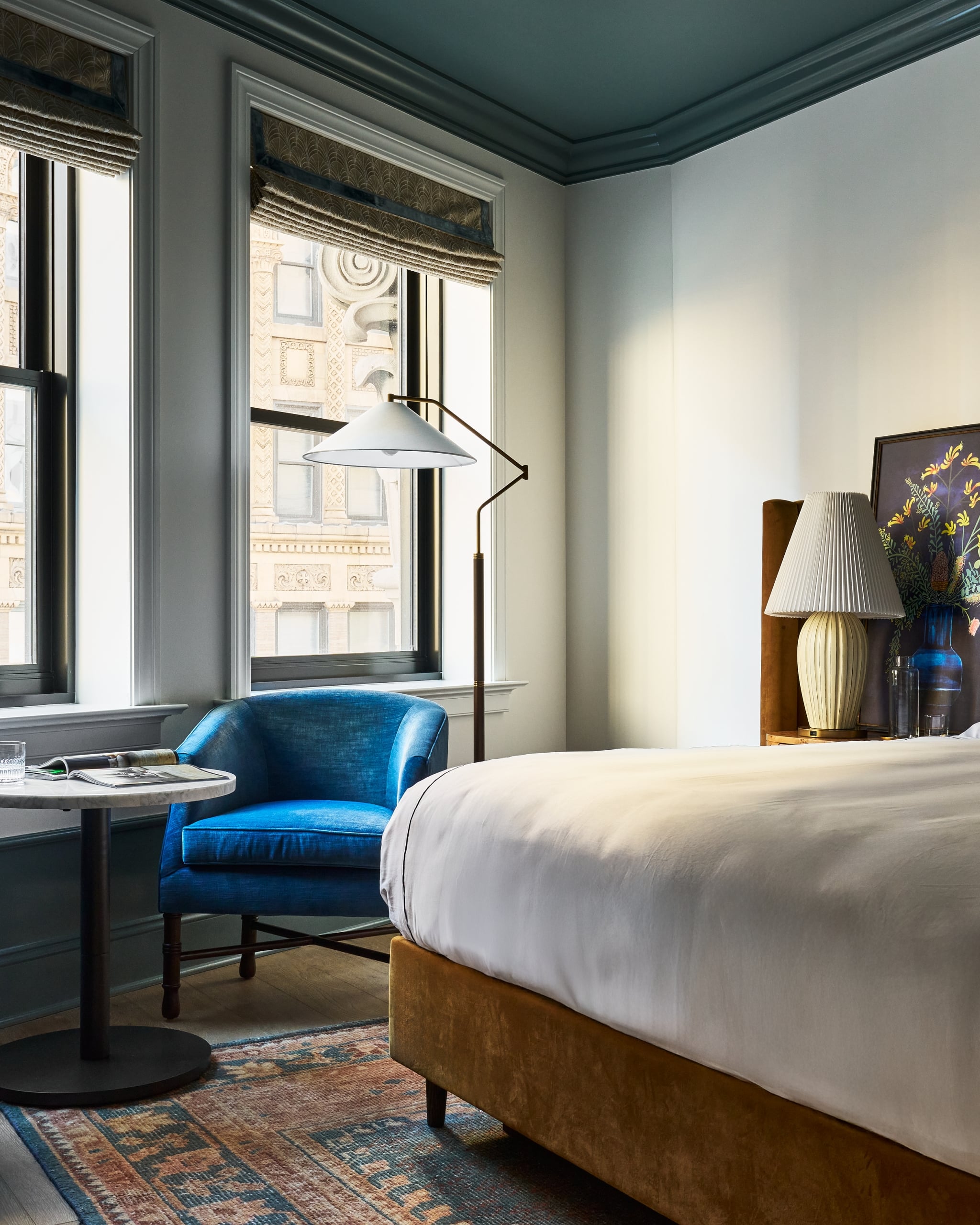 A plush blue armchair sits by tall windows overlooking ornate city buildings. A bed with white sheets and a patterned rug furnish the elegant room.