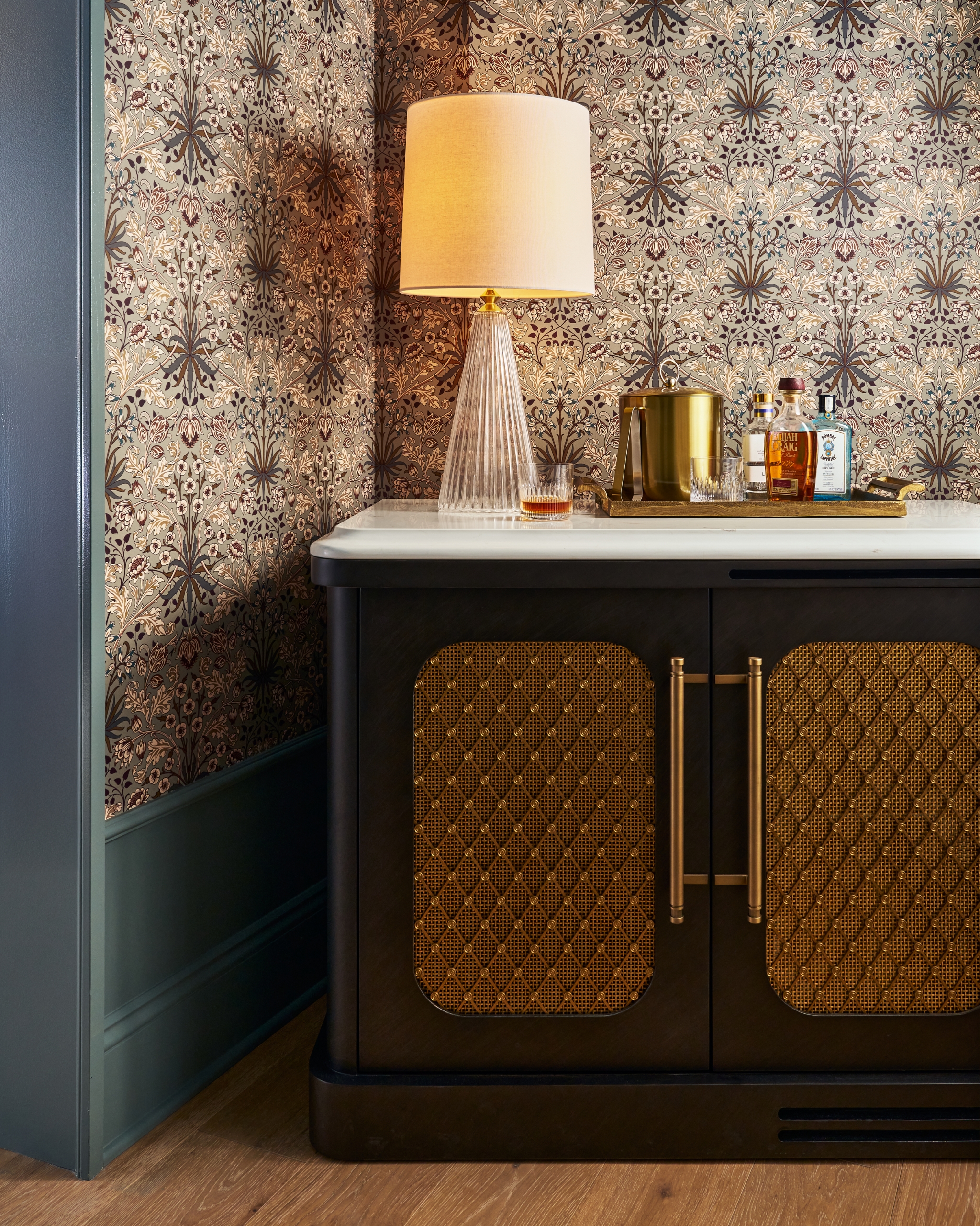 A black bar cabinet with gold patterned doors displays a lamp and a tray holding drinks, set against floral wallpaper and dark green wainscoting. Text: ELIJAH CRAIG Small Batch KENTUCKY STRAIGHT BOURBON WHISKEY 94 PROOF, BOMBAY SAPPHIRE London Dry GIN.