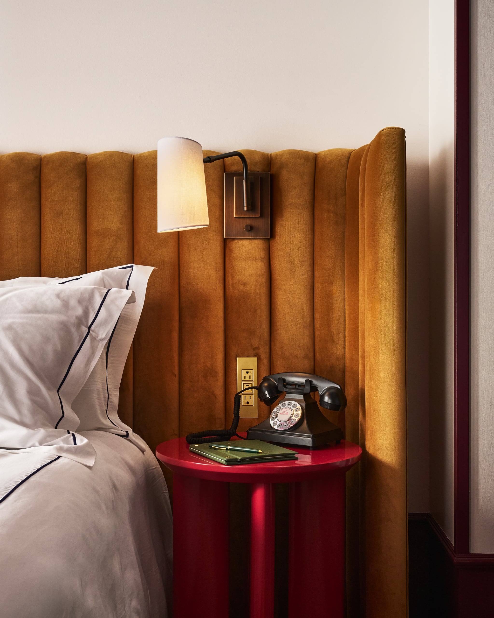 A black rotary phone sits on a red nightstand beside a bed with white linen and an amber velvet headboard, illuminated by a wall sconce. Text on phone dial: "WESTERN ELECTRIC PROPERTY OF THE BELL SYSTEM 500 AUG 58"