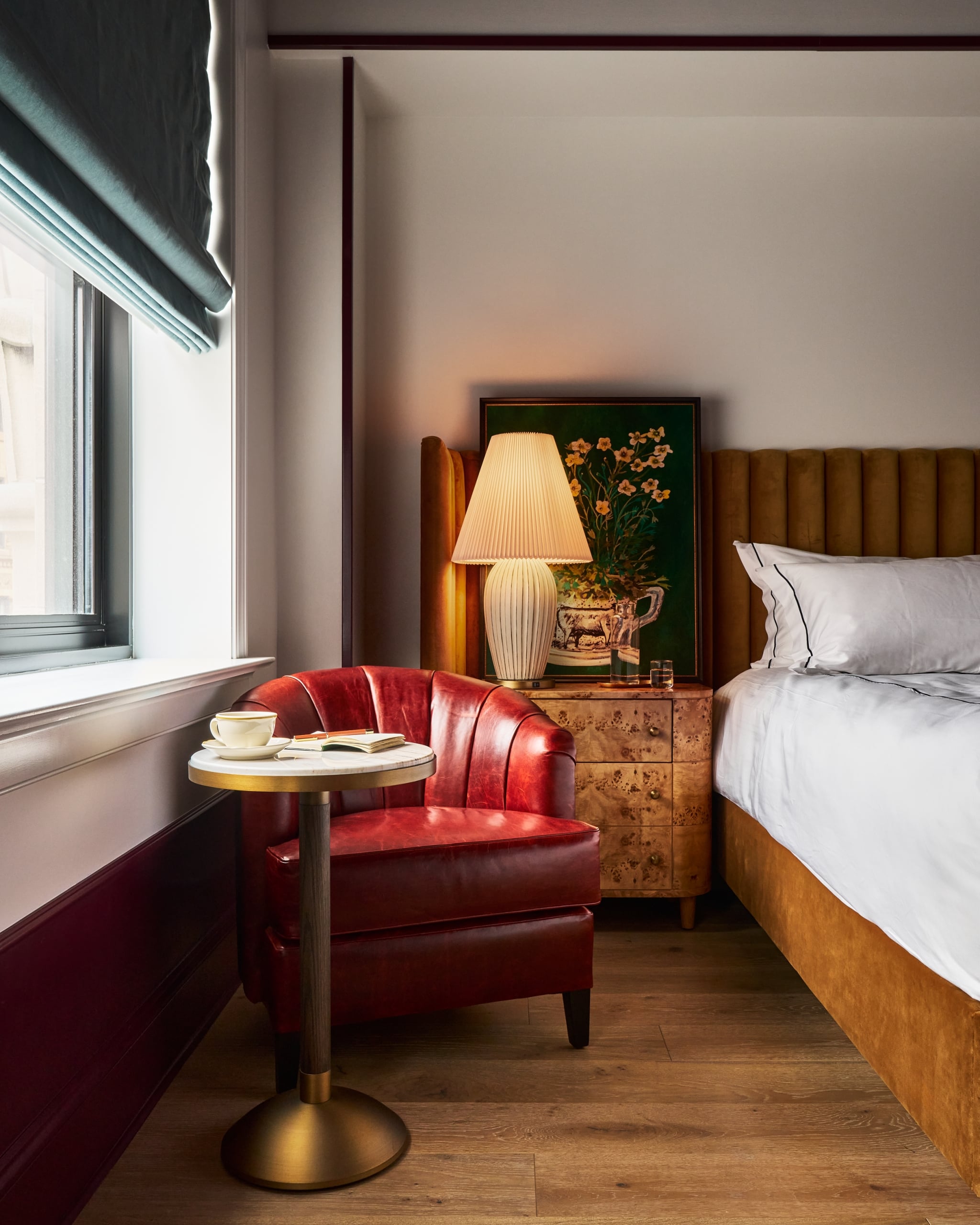 A red leather armchair sits beside a window with a blue blind, next to a marble-top table holding a cup and open book, in a warm-toned bedroom.
