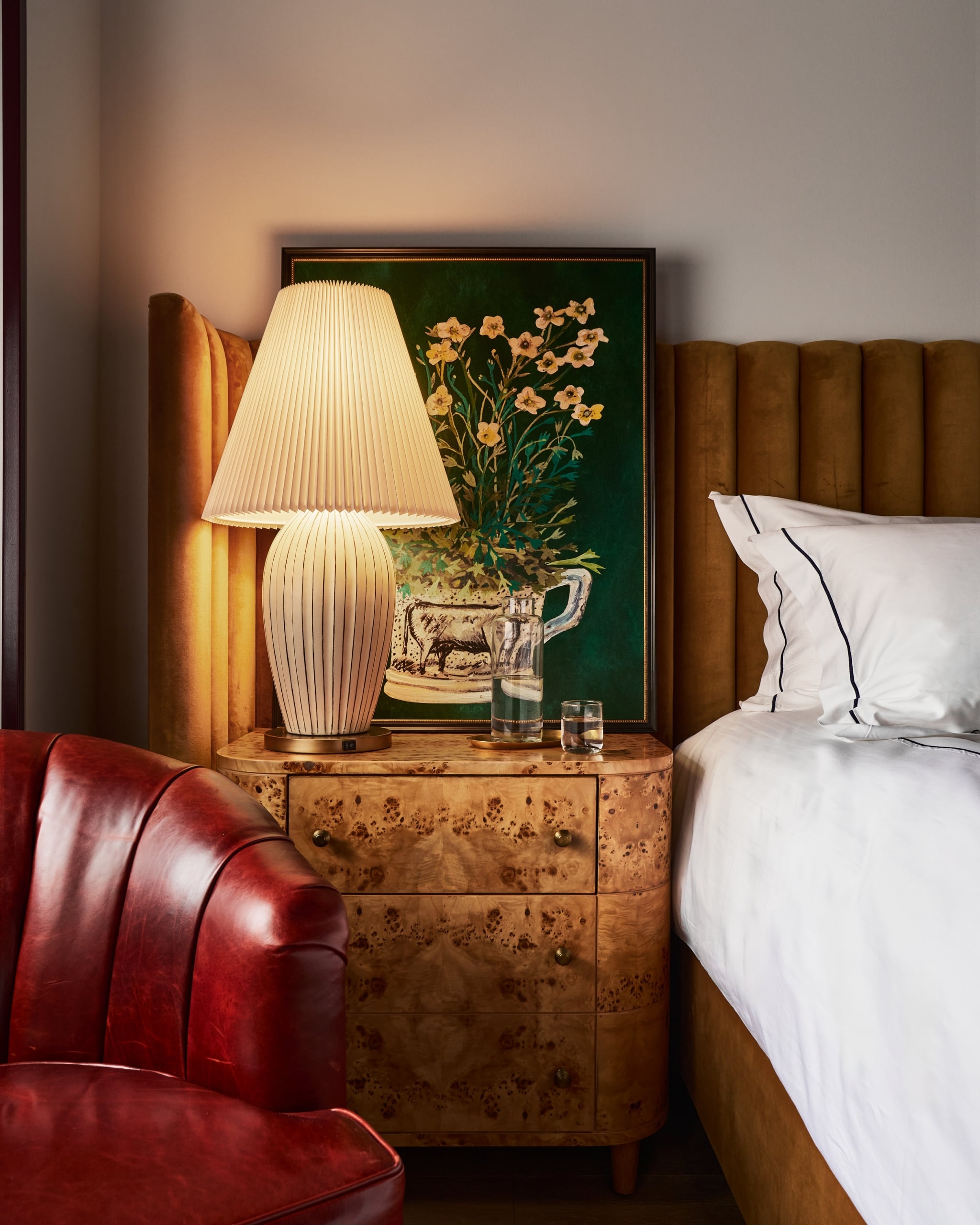 A wooden nightstand holds a lit pleated lamp, a floral painting, and water. It sits between a red leather armchair and a bed with a brown velvet headboard.