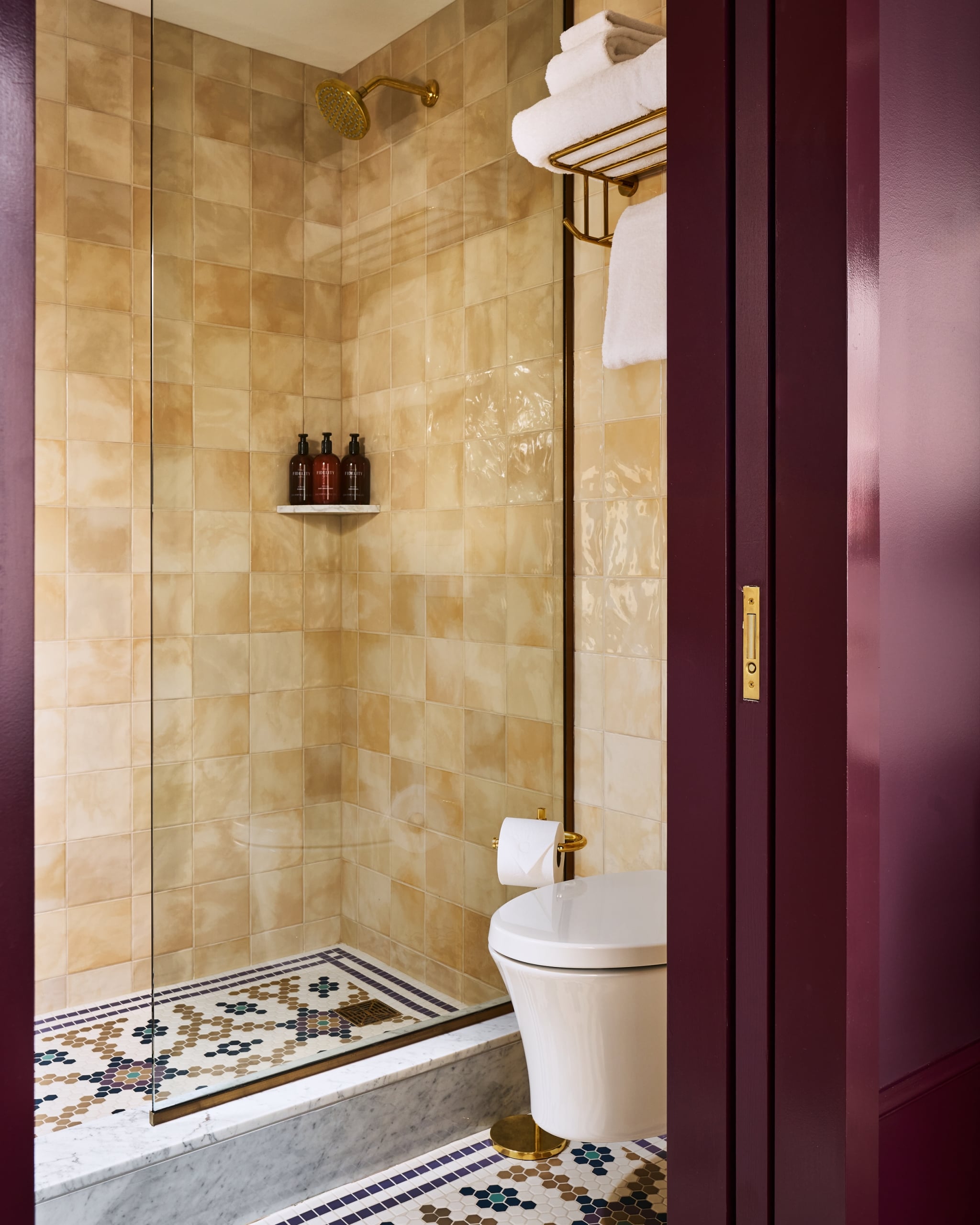 A shower stall features a gold head and three brown bottles, next to a white toilet. Beige tiled walls, a mosaic floor, and a burgundy doorway complete the scene.