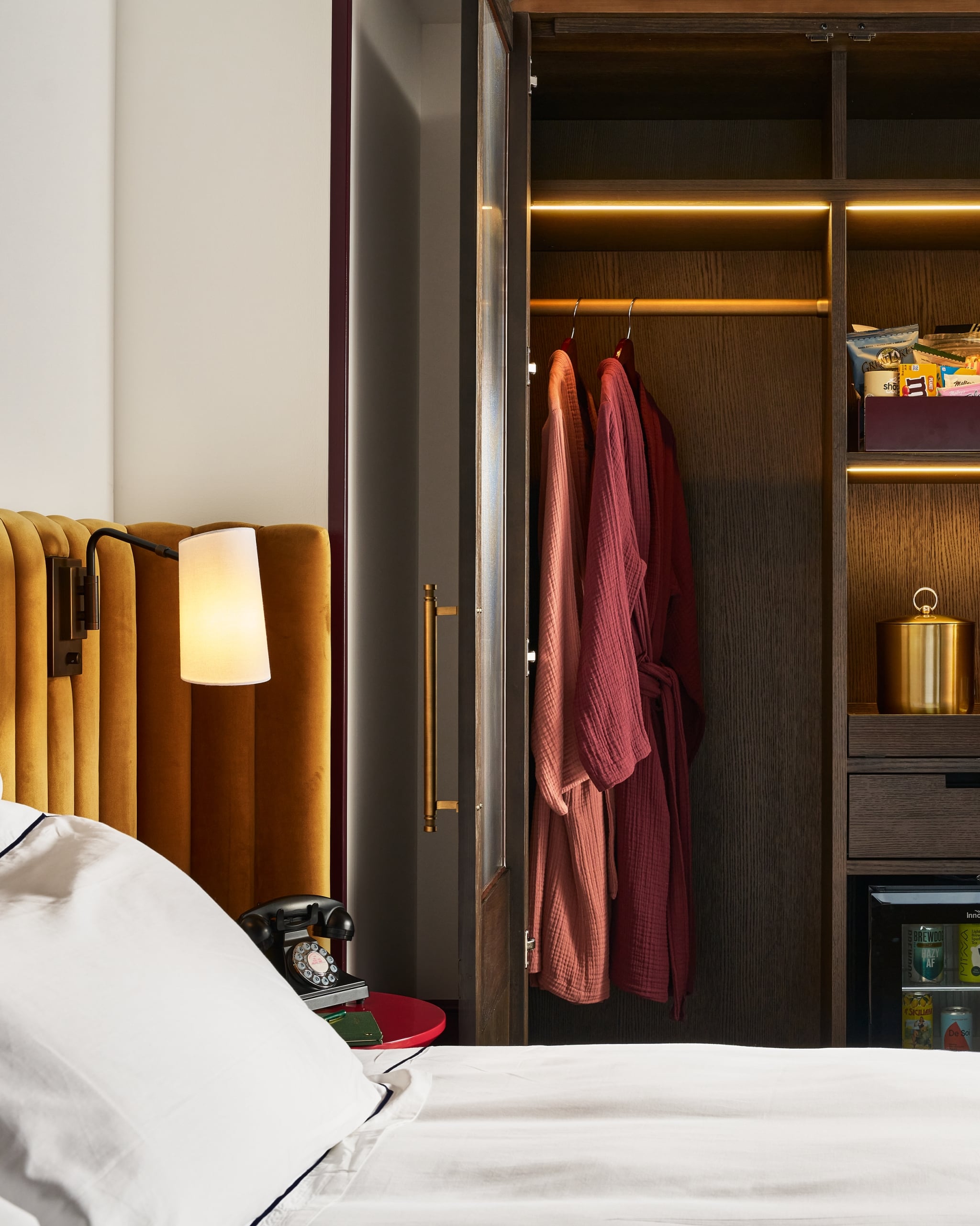 A glowing bedside lamp illuminates a bed with a mustard headboard next to an open dark wood wardrobe. It holds two pink robes, snacks ("GREAT LAKE", "sho", "Maltesers"), and a minibar ("BREW DOG", "HAZY AF", "SKILLIWAN", "De Sol", "Inno").