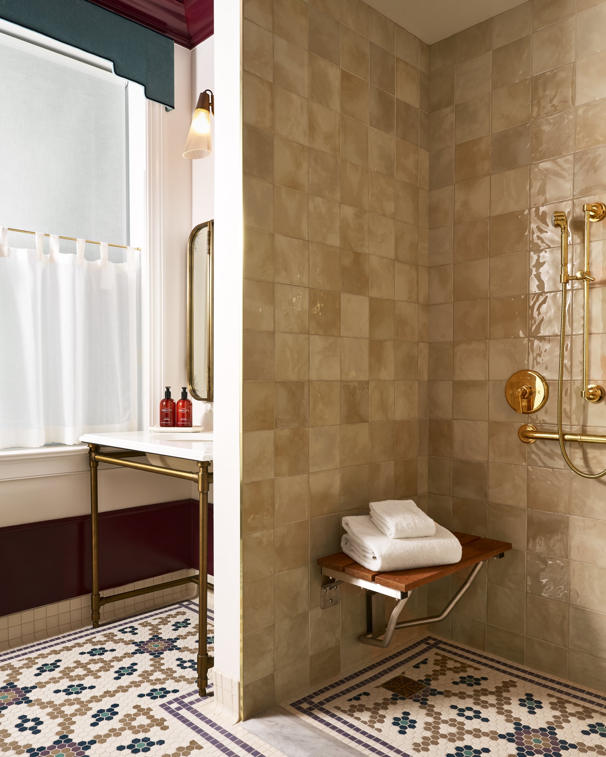 Tiled bathroom featuring a white vanity with two bottles: "FIDELITY'S HAND SOAP & LOTION 10 FL OZ (300 ML)". Adjacent shower boasts gold fixtures, a wooden bench, and folded white towels.