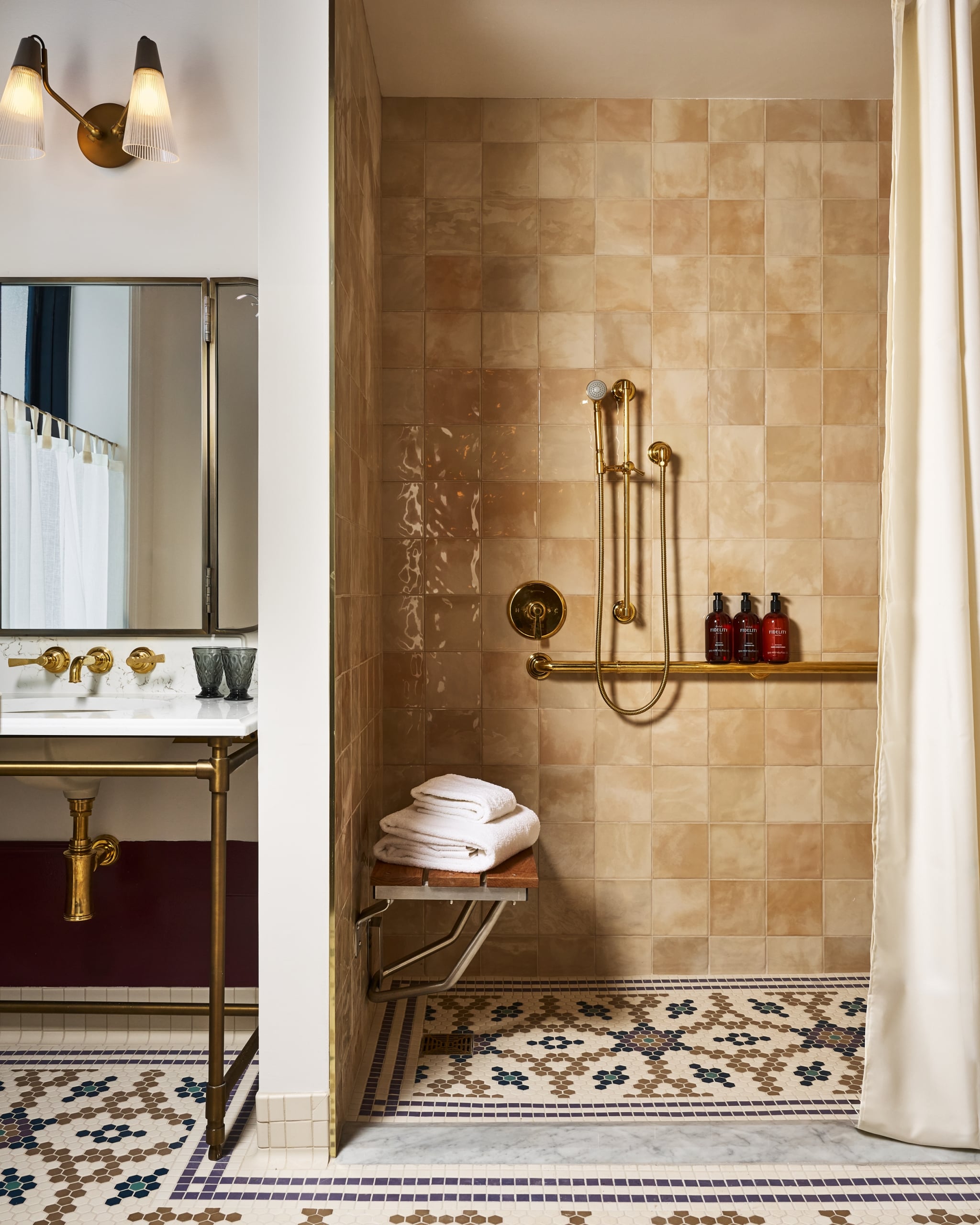 A brown-tiled shower with brass fixtures includes a wooden bench holding white towels. The patterned floor continues past a marble vanity. Three bottles read: 'FIDELITY HOTEL GOOD PEOPLE GOOD TIMES e 500 ml e 16.9 fl.oz.'