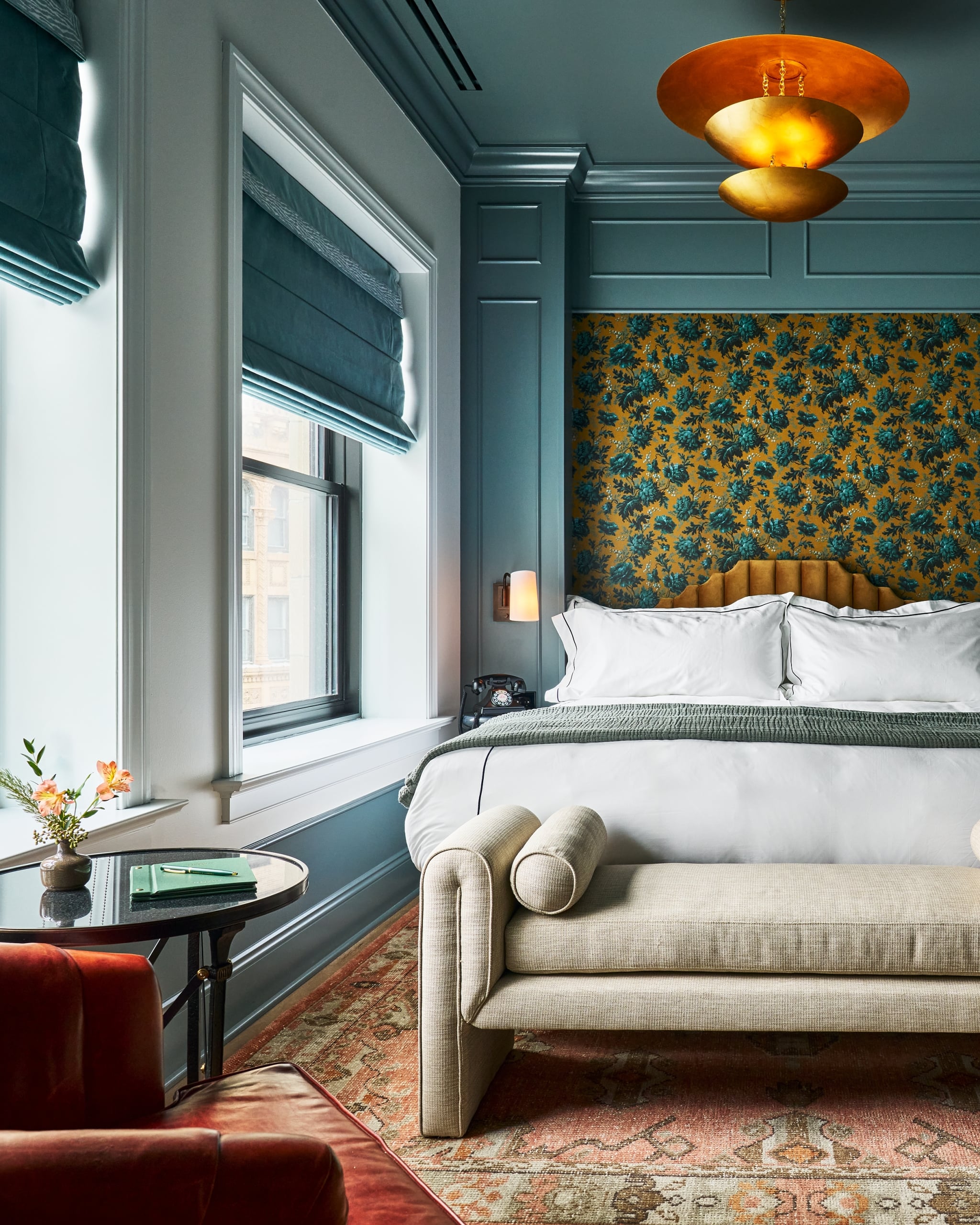 Neatly made bed with white bedding sits in a luxurious room. It features a mustard floral wallpaper accent, teal paneling, a large window, and a striking gold tiered chandelier.