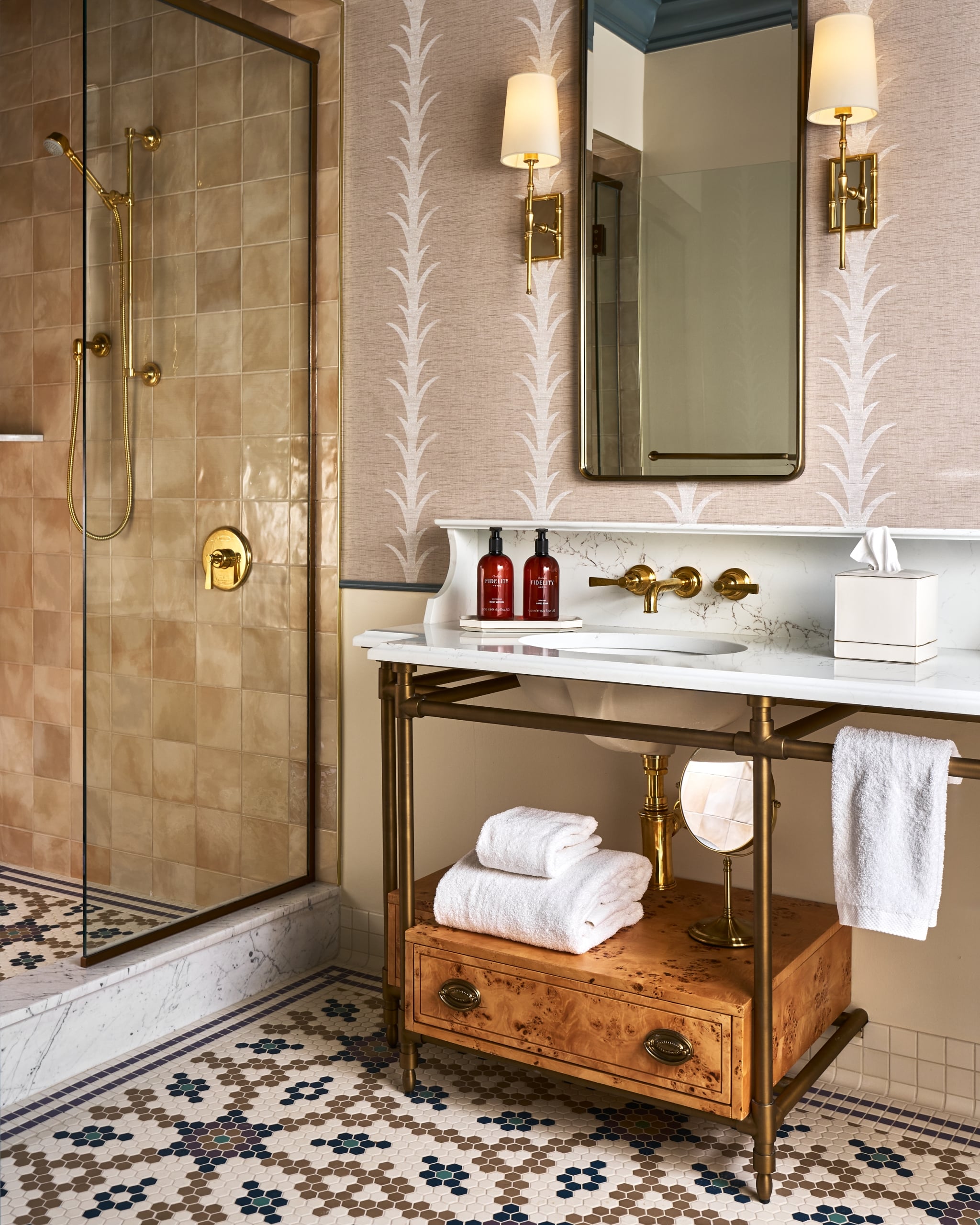 A modern bathroom vanity, with a marble top and sink, stands next to a glass-enclosed shower stall. The room features patterned wallpaper and mosaic floor tiles, accented with gold fixtures.