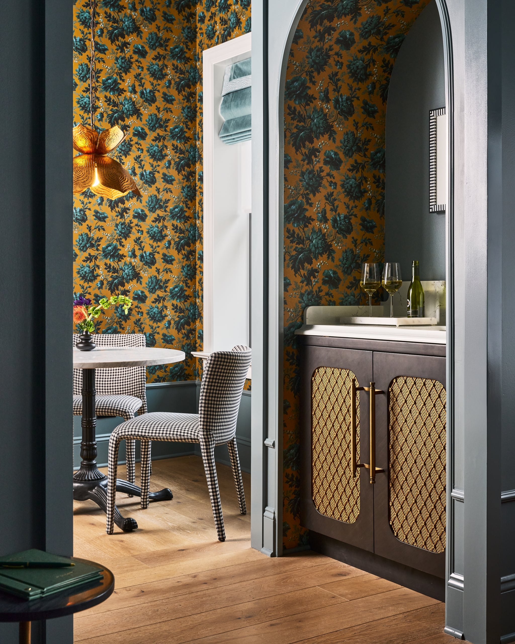 Dining nook with a marble table and houndstooth chairs, and a bar cabinet with wine glasses, are framed by an archway. Both spaces feature mustard yellow floral wallpaper.