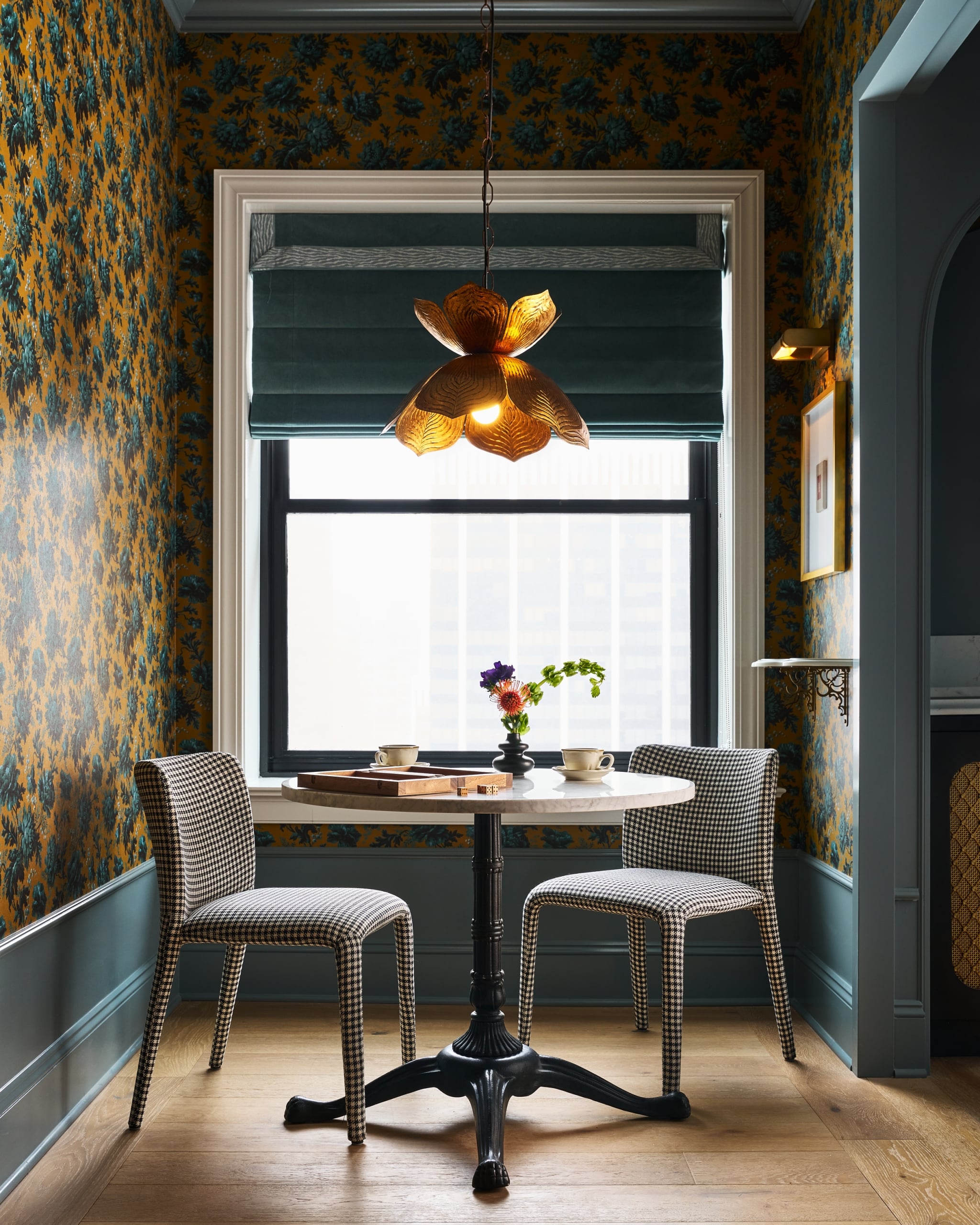 A round marble table and two houndstooth chairs sit by a window. A game, dice, cups, and flowers adorn the table in a gold floral wallpapered room.