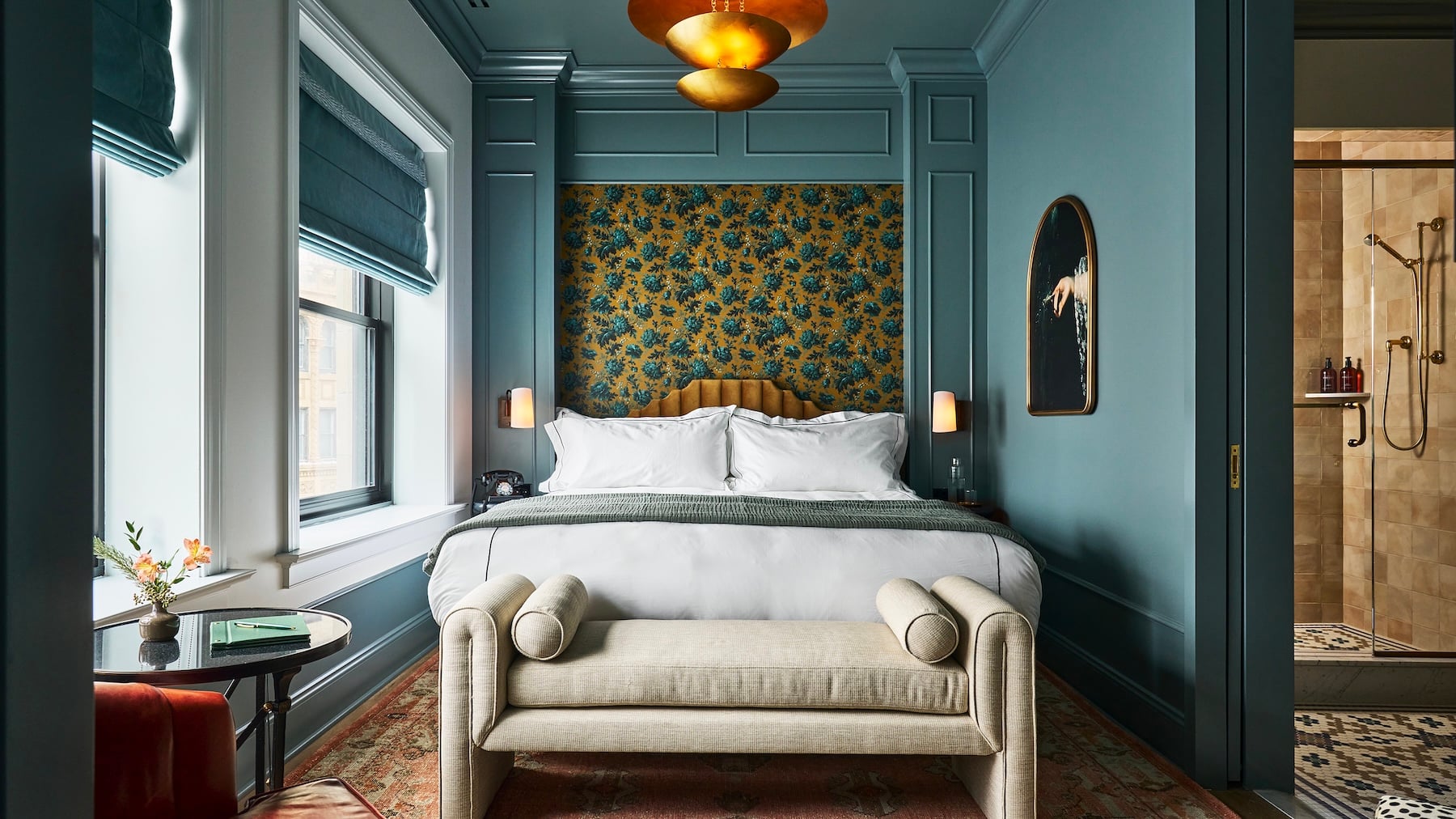 A plush white bed with a padded gold headboard sits against floral wallpaper. Teal paneled walls frame the room, with a shower area visible nearby.