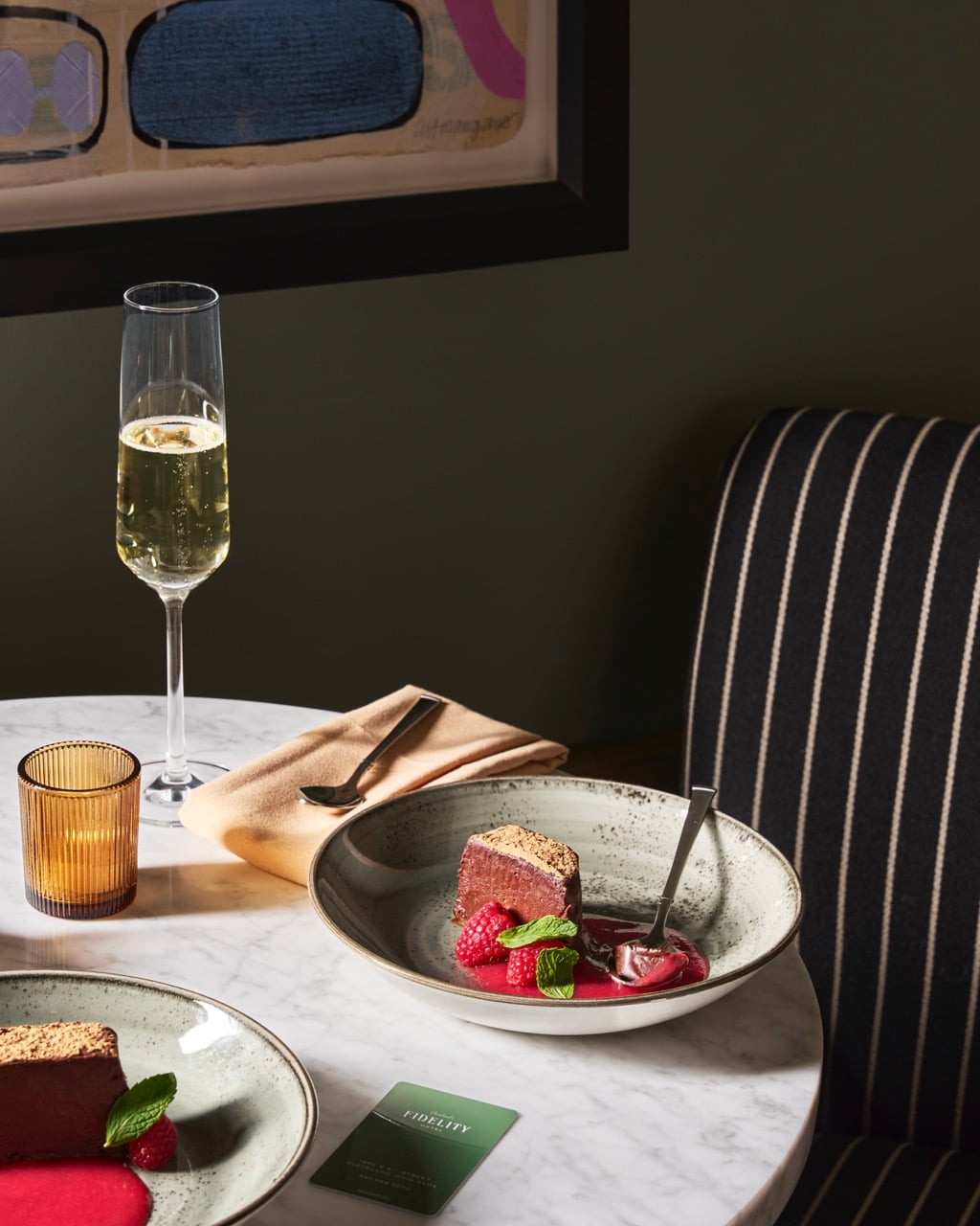 Two chocolate desserts with raspberries and mint sit on a marble table with champagne. A "FIDELITY" card, napkin, and candle holder complete the setting against a dark wall.