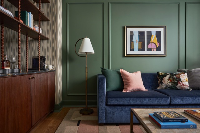 Blue velvet sofa and colorful pillows against a dark green paneled wall with abstract art. A floor lamp stands by a wooden bar cabinet. Coffee table books: "PHOTOGRAPHY", "CINEMATOGRAPHY".