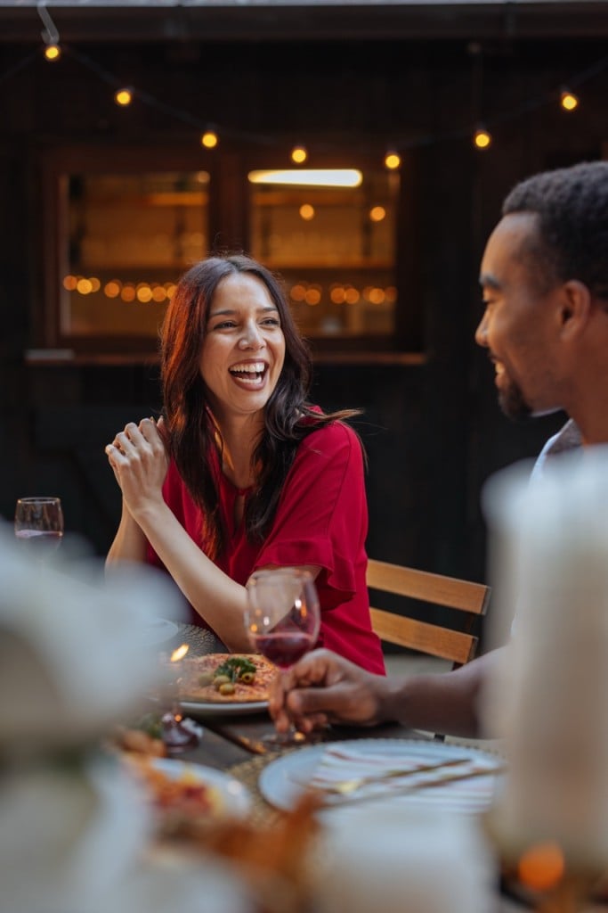 A woman in a red shirt laughs heartily, hands clasped, at an outdoor dinner table with a man, under string lights in the evening.