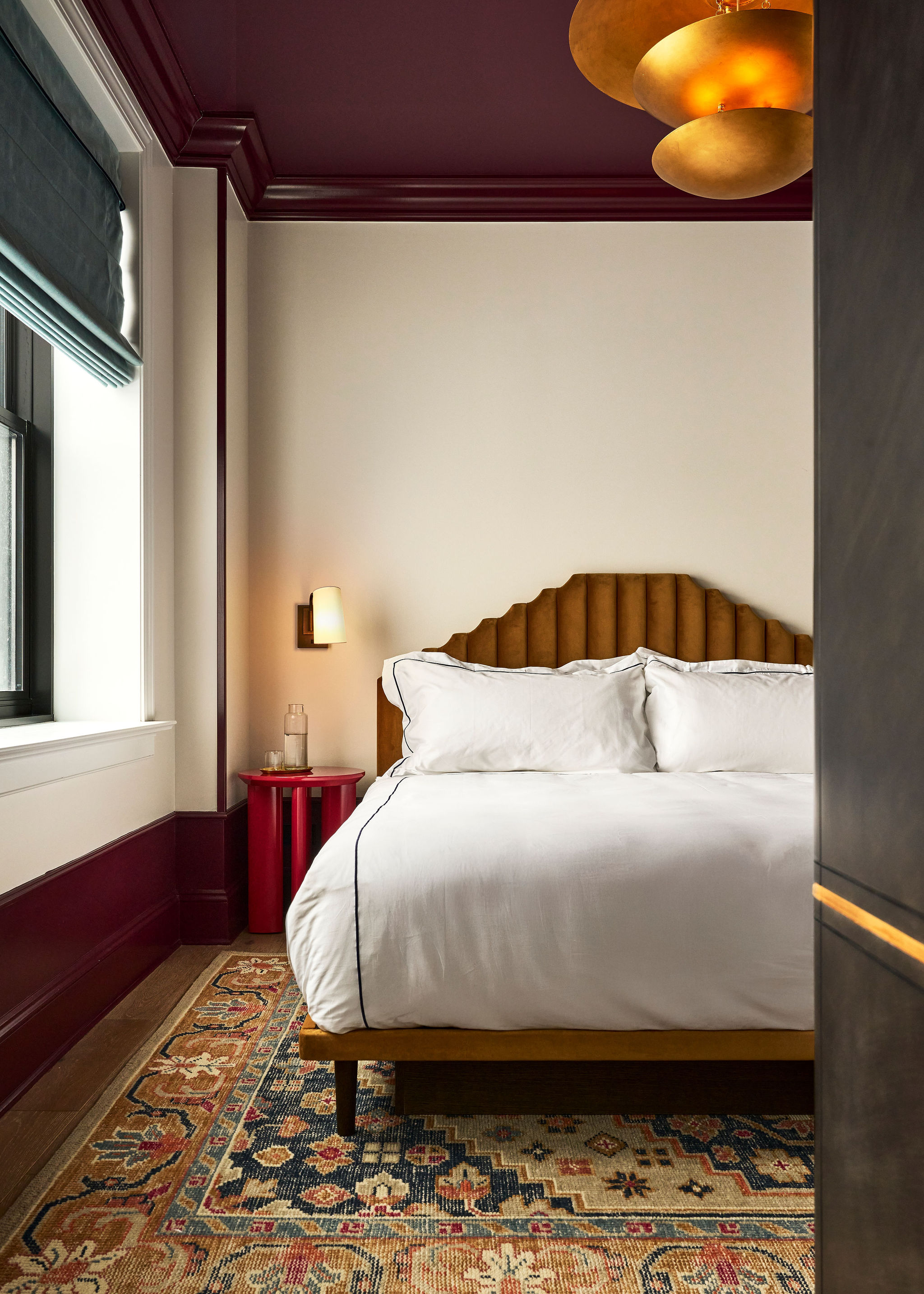 A bed with white linens and a brown headboard is positioned against a wall, with a red nightstand and lamp beside it.