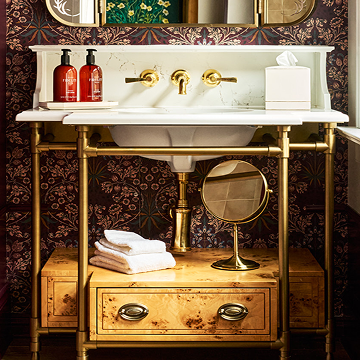 A brass-framed vanity features a white marble top, undermount sink, and gold wall taps. Against dark floral wallpaper, it displays toiletries, including two red bottles labeled "FIG+ROUGE HAND WASH" and "FIG+ROUGE HAND LOTION."