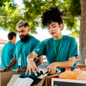 A woman with curly hair sorts clothes into a cardboard box, outdoors with a bearded man volunteering next to her and bottles of orange liquid on a table.
