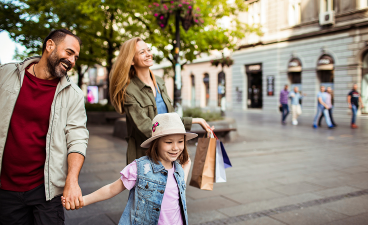 A happy family of three, a man, woman, and young girl, smiling while walking hand-in-hand with shopping bags down an urban street.