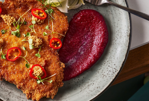 Crispy breaded cutlet garnished with chili, mustard seeds, and microgreens, served alongside beet puree and a fennel salad with a fork, on a speckled gray plate.