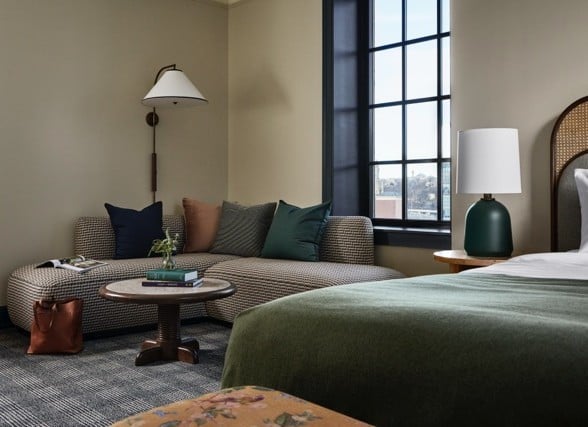 A modern hotel room is furnished with a patterned curved sofa, coffee table, and bed with a green blanket and woven headboard, beside a large window.