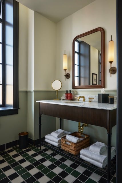 A dark wood vanity with a marble sink, golden faucet, and mirror is flanked by sconces. White towels are neatly stacked below, set in a bathroom with checkered floor tiles.