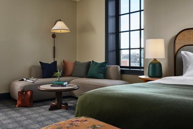 An L-shaped sofa with pillows and a round coffee table sits by a window in a modern hotel room, with a bed and a green blanket visible on the right.