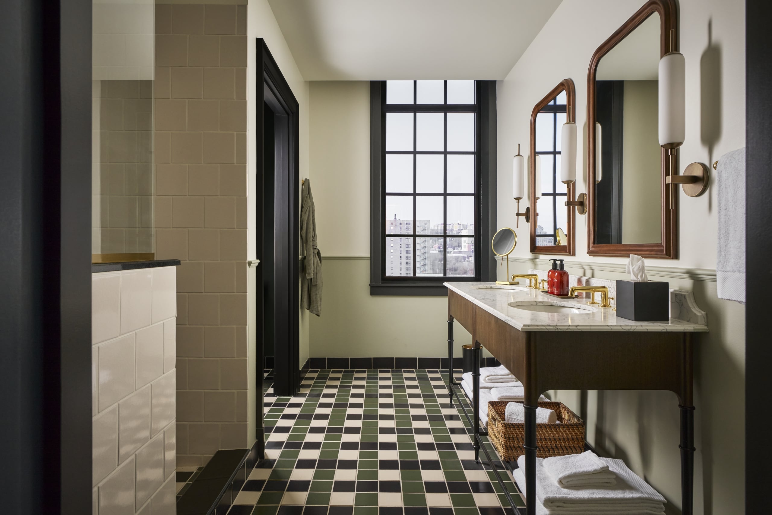 A modern bathroom showcases a double vanity, twin framed mirrors, and a large grid window overlooking a city. It features a black, white, and green checkered floor and tiled shower walls. MALIN+GOETZ body wash.