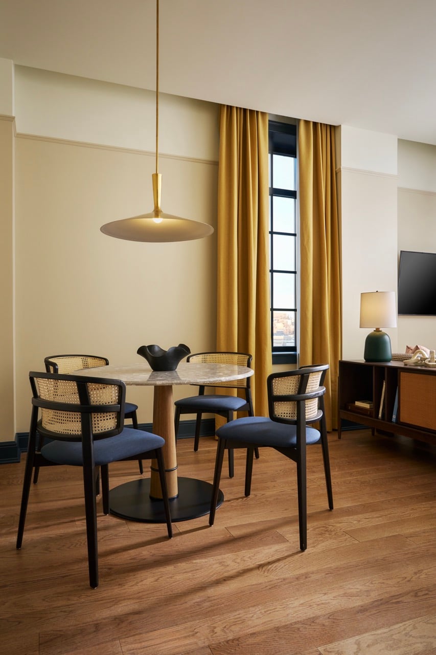 A round marble dining table with four dark cane-back chairs, arranged beneath a brass saucer pendant light, in a room with yellow curtains, beige walls, and wood flooring.