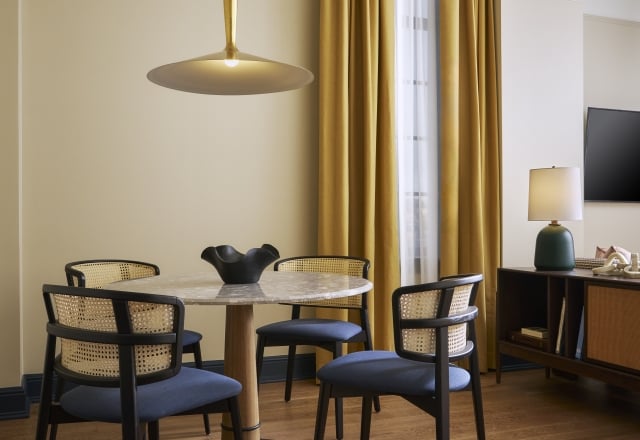 A round marble dining table and four dark chairs with cane backs and blue cushions sit under a brass pendant lamp. Pale yellow walls, wooden floor, and yellow curtains complete the room.