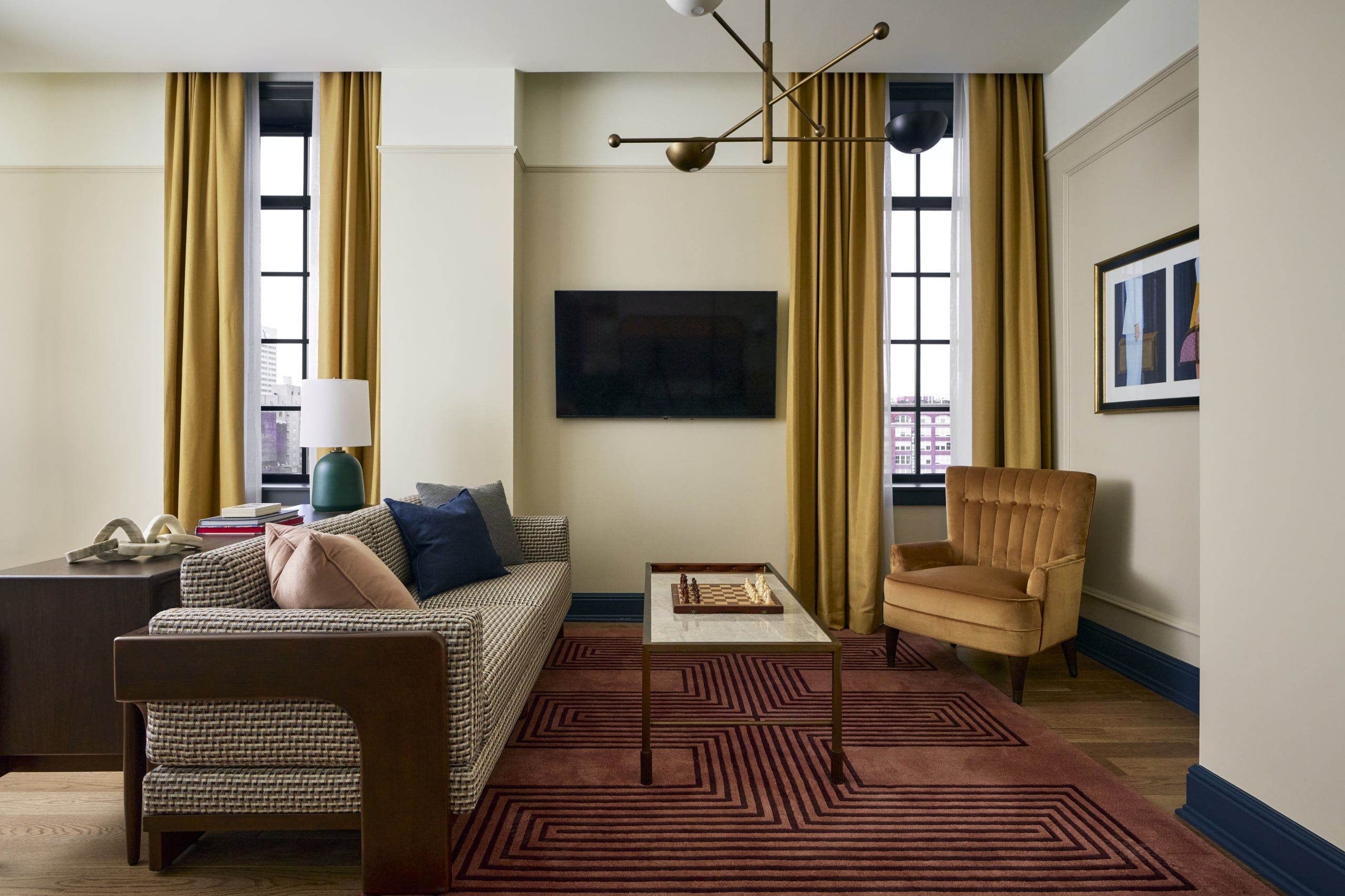 A modern living room features a patterned sofa, chess-set coffee table, and gold armchair, all facing a wall-mounted TV and tall windows in a sophisticated interior.