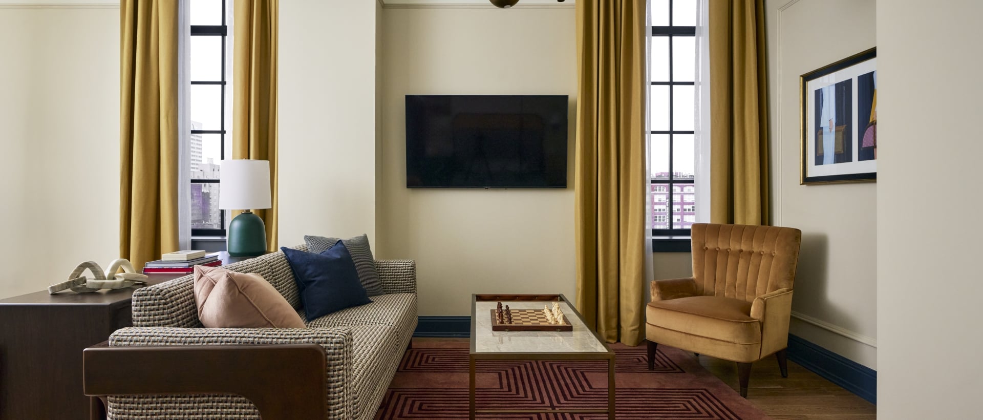 A checkered sofa and velvet armchair frame a coffee table with a chess set, furnishing a modern lounge. A TV is mounted opposite, with mustard curtains flanking large windows.