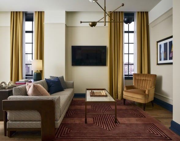 A modern living room features a sofa, coffee table with a chessboard, and an armchair, illuminated by windows with golden curtains and a stylish chandelier.