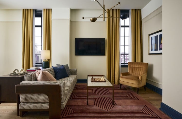 A modern living room features a sofa, coffee table with a chessboard, and an armchair, illuminated by windows with golden curtains and a stylish chandelier.